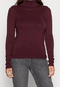 Kobieta w dopasowanym bordowym swetrze z golfem i szarych dżinsach, stojąca na tle jasnej, jednolitej powierzchni.