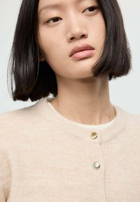 Cardigan bej deschis cu guler rotund, având capse metalice mici, textură moale și un design ajustat. Fundalul este neutru.