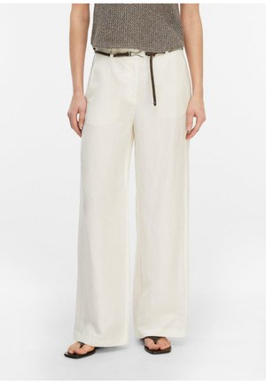 Frau trägt hoch taillierte, weit geschnittene cremefarbene Hose mit braunem geflochtenem Gürtel, gestricktes ärmelloses Oberteil und schwarze Zehensandalen.