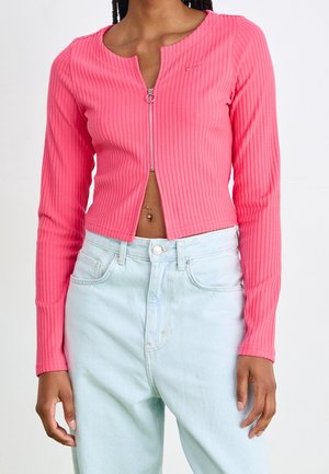 Personne portant un haut court côtelé rose à manches longues avec une fermeture éclair à l'avant et un jean bleu clair taille haute.