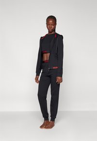 Zwarte cropped hoodie met rode accenten en logo, gecombineerd met zwarte joggers met een bijpassende logo tailleband. Gladde stof en aansluitend ontwerp.
