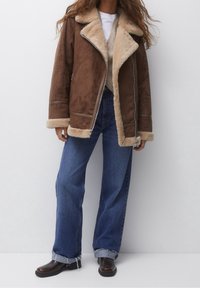 Brun ruskinds shearling jakke med en tan farvet pelsforing, brede klapper og lynlåslukning. Kombineret med blå jeans med lige ben og brune ankelstøvler.