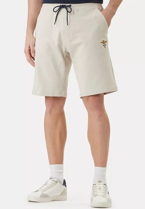 Uomo che indossa pantaloncini beige con coulisse e logo, abbinati a calzini bianchi e scarpe da ginnastica bianche, in piedi su uno sfondo semplice.