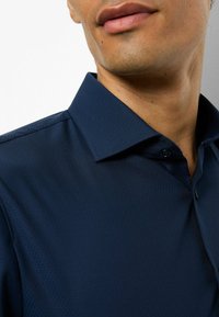 Marineblaues texturiertes Hemd mit spitzem Kragen, das ein dezentes geometrisches Muster und einen einzelnen Knopf am Kragen aufweist. Weicher Stoff.