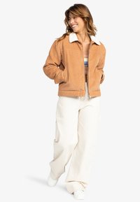 Tan corduroy jas met witte nepbont kraag, vooraan met rits, twee zijzakken, gecombineerd met lichtgekleurde wijde broeken en witte sneakers.