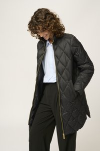 Zwarte gewatteerde jas met een gouden rits, hoge kraag en lange mouwen. Draagt over een lichtblauw overhemd met knopen en zwarte broek.