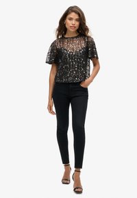 Zwarte zich verglanzende crop top met korte mouwen, met een doorschijnende overlay en patroon details, gedragen met strakke zwarte jeans en sandalen.