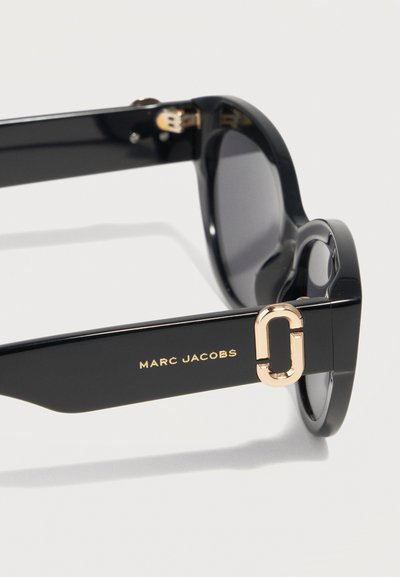 Μαύρα γυαλιά ηλίου Marc Jacobs με παχιά πλαίσια, σκούρους φακούς και χρυσό λογότυπο και διακοσμητικά στοιχεία στα πλαϊνά μέρη.