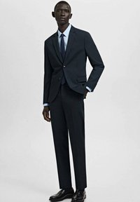 Mężczyzna ubrany w ciemnogranatowy garnitur, jasnoniebieską koszulę, granatowy krawat i czarne eleganckie buty, stojący z jedną ręką w kieszeni.