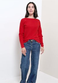 Maglione a costine rosso con maniche lunghe, indossato su jeans in denim blu. Il maglione presenta un colletto rotondo e una vestibilità comoda.