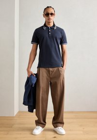 Námornícky polo tričko s bielymi akcentmi, béžové široké nohavice, drží námornícku mikinu a nosí slnečné okuliare s minimalistickým dizajnom.