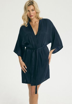 Blondínka nosi tmavomodrý saténový župan so širokými rukávmi a zviazaným opaskom, stojaca pred jednoduchým svetlým pozadím.