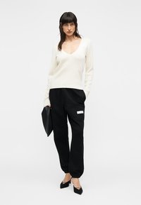 Κρεμ πουλόβερ με v-λαιμόκοψη και μακριά μανίκια, σε συνδυασμό με μαύρο ανοιχτό τζόγκερ με ετικέτα στο πλάι. Μαύρο τσάντα φάκελος και μυτερά μοκασίνια.