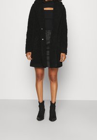 Manteau teddy surdimensionné noir avec une finition texturée, doté d'une capuche, d'une fermeture à boutons et de poches latérales, associé à un haut noir ajusté et une jupe en cuir.