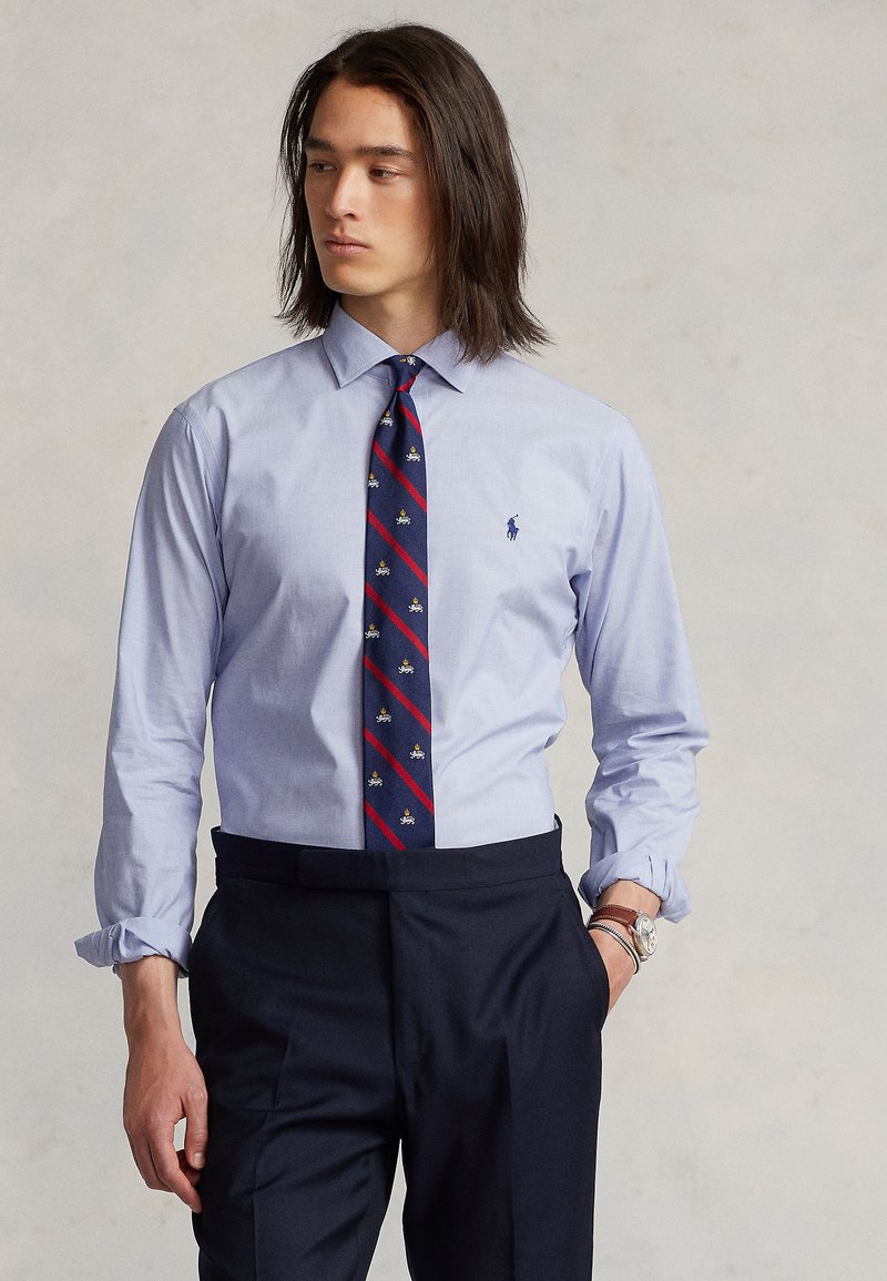 Lyseblå skjorte med lange ermer og krage, kombinert med et marineblått slips med røde striper og små broderte motiver, brukt sammen med marineblå bukser.