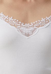 Débardeur blanc avec un col en V et une délicate bordure en dentelle blanche, présentant des motifs de papillons le long du col, texture côtelée sur l'ensemble.