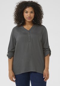 Blouse légère gris foncé avec un décolleté en V, patte de boutonnage et manches roulées, fabriquée dans un tissu lisse. Présente une coupe décontractée.