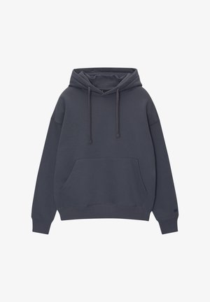 Persona vistiendo una sudadera con capucha de color gris oscuro, pantalones sueltos color crema y zapatillas marrones, de pie con las manos en los bolsillos de la sudadera.