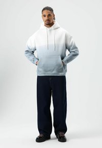 Pullover hoodie in een verloop van wit naar lichtblauw stof, met een zak aan de voorkant en een capuchon met trekkoord. Gecombineerd met losse donkere jeans en zwarte schoenen.