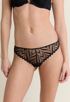 Frau trägt schwarze Spitzenunterwäsche mit Blätter-muster Netzpanties und passendem BH, steht vor neutralem Hintergrund.