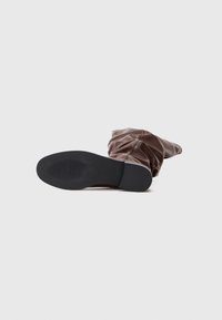 Bota marrón, holgada hasta la rodilla, con material suave, punta redondeada y suela de goma negra con rayas horizontales para mayor agarre.