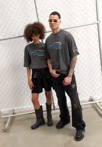 To modeller bærer løstsiddende charcoal t-shirts med blå grafisk tekst. Den mandlige model har slidte sorte jeans og chunky sneakers. Den kvindelige model sætter sin t-shirt sammen med sorte shorts og knæhøje, nittede støvler.