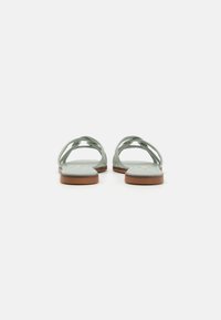 Sandales plates vertes avec des sangles entrelacées, semelles plates et une base marron, présentant une surface lisse et un design minimaliste.