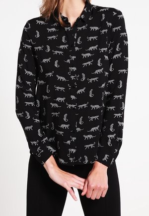 Chemise noire à manches longues boutonnée avec motif léopard blanc portée par une personne les mains jointes devant.