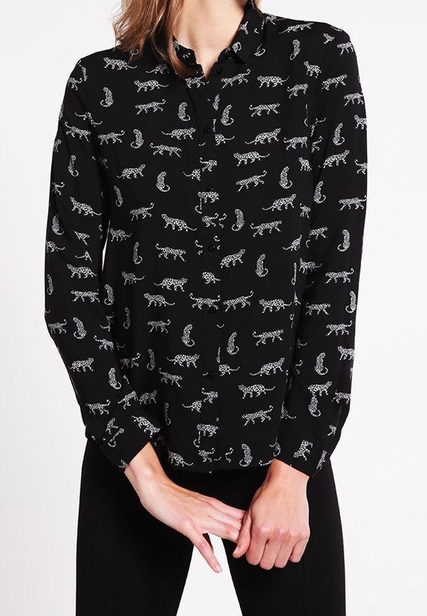 Chemise noire à manches longues boutonnée avec motif léopard blanc portée par une personne les mains jointes devant.