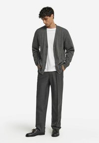 Młody mężczyzna ubrany w ciemnoszary zapinany kardigan, białą koszulkę, szare spodnie i czarne buty, stojący z rękami w kieszeniach, patrzący w dół.