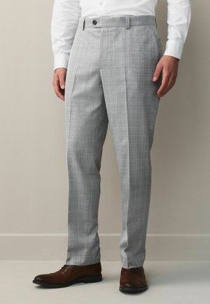 Man wearing light gray plaid dress pants, white shirt, and brown leather dress shoes, standing against a plain beige wall.