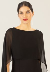 Schwarzes Kleid mit U-Boot-Ausschnitt und durchsichtigen Cape-Ärmeln. Glatte Textur, elegante Silhouette, ergänzt durch hängende Ohrringe.