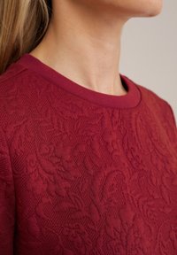 Burgunderfarbener, strukturierter Oberteil mit floralem Muster und Rundhalsausschnitt. Der Stoff wirkt weich mit einem gestrickten Design.