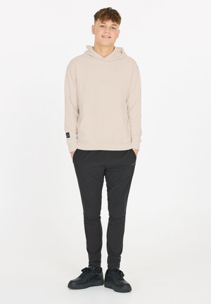 Jeune homme debout, mains dans les poches, portant un sweat à capuche beige, un pantalon noir slim et des baskets noires, sur un fond blanc uni.