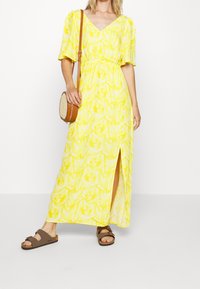 Robe maxi florale jaune avec des manches bouffantes courtes, un col en V et une fente sur le côté. Portée avec des sandales à glisser brunes et un petit sac à main rond.