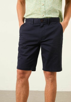 Man wearing navy blue casual shorts and a light green patterned short-sleeve shirt, with hands in pockets, standing indoors.