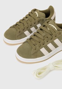 Baskets en suède vert olive avec des rayures blanches, lacets plats, partie supérieure texturée et semelle extérieure en caoutchouc naturel. Comprend un lacet supplémentaire d'un blanc cassé.