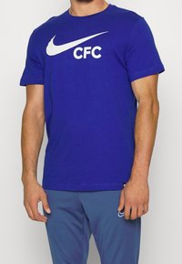 Niebieski t-shirt z krótkim rękawem wykonany z bawełny, ozdobiony dużym białym logo Nike w kształcie znaku „swoosh” oraz nadrukiem „CFC” z przodu.