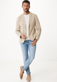 Beige leichte Blazer über einem weißen T-Shirt, kombiniert mit hellblauen Jeans und beigen Slippern, vor einem neutralen Hintergrund.