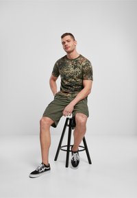 T-shirt a maniche corte in camouflage nei toni di verde e marrone, abbinato a pantaloni corti color oliva. Sneakers bianche e nere. Seduto su uno sgabello nero.