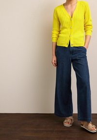 Cardigan giallo a coste con scollo a V e tre bottoni, abbinato a pantaloni in denim scuro a gamba larga e sandali con stampa animale.