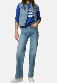 Mujer vestida con un chaleco de mezclilla azul claro y jeans, sudadera azul con texto blanco, y zapatillas blancas, de pie con una mano en el bolsillo.