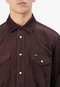 Női torsó sötét bordó corduroy Wrangler ingben, nyomógombokkal és két mellzsebbel, az egyik zsebében Wrangler logós címkével.