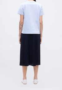 Personne portant une chemise bleu clair à manches courtes, une jupe midi plissée noire, des chaussures blanches, debout avec les bras détendus, vue de dos.