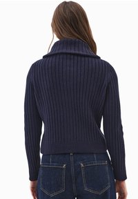 Meriharmaa ribattu neule, jossa on korkea kaulus, istuva muotoilu ja pitkät hihat. Materiaali on pehmeä, yhdistettynä tummansinisiin farkkuihin.