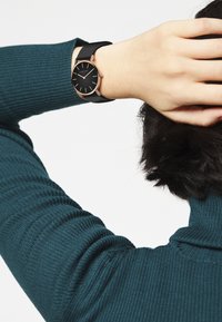 Zwarte horloge met een ronde wijzerplaat, roségouden accenten en een zwarte siliconen band, gedragen om een pols bedekt met een geribbelde teal trui.