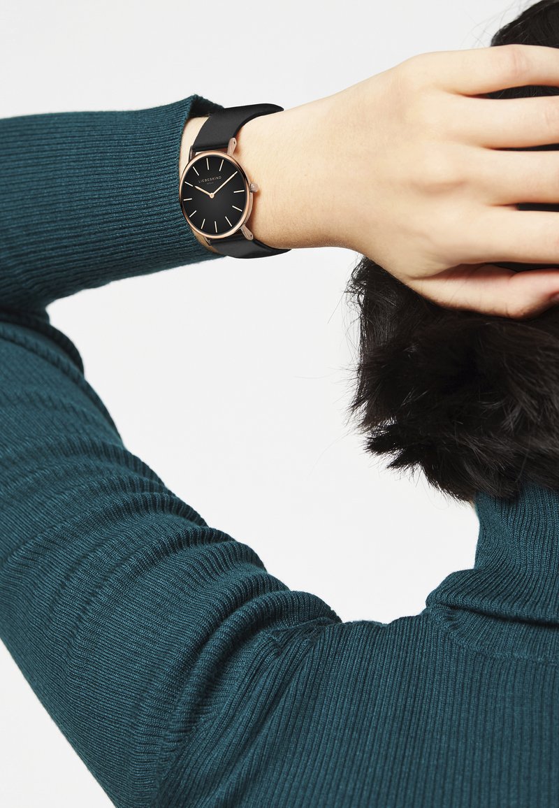 Zwarte horloge met een ronde wijzerplaat, roségouden accenten en een zwarte siliconen band, gedragen om een pols bedekt met een geribbelde teal trui.