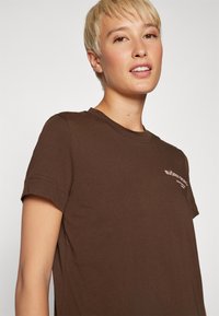 T-shirt in cotone marrone con maniche arrotolate e collo a giro. Presenta un piccolo logo bianco sul lato sinistro. Texture morbida e vestibilità casual.