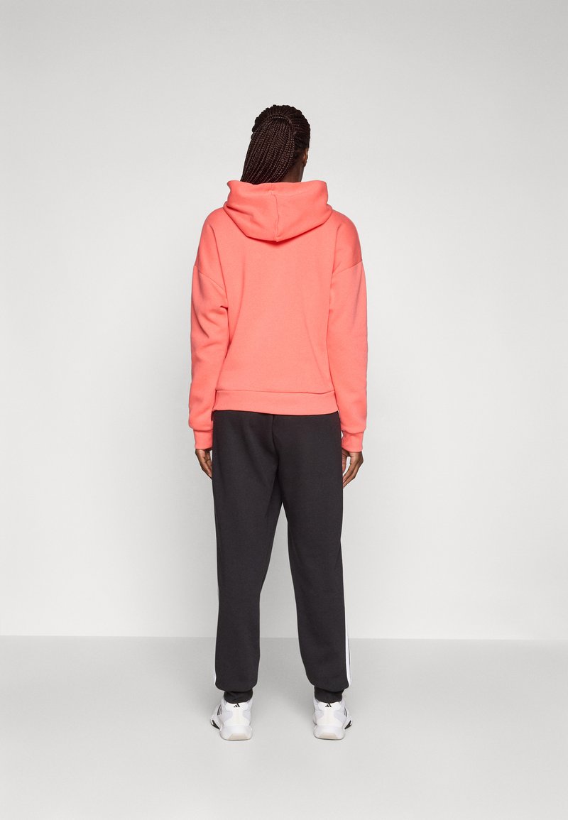 Sudadera de color coral con cuello alto y dobladillo acanalado, combinada con pantalones de chándal gris oscuro que tienen franjas blancas a los lados. Textura suave, estilo deportivo.