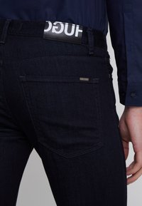 Tmavě modré džíny s přiléhavým střihem, černá kožená visačka s bílým logem na pase a decentní detaily na zadních kapsách.